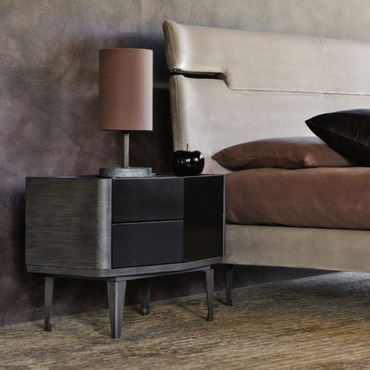 A stylish bedroom featuring a beige upholstered bed with brown bedding. Beside the bed is a *Modern Bedside Cabinet With Two Drawers*, designed with sleek dark wood and black drawer fronts, supporting a contemporary brown table lamp and a small black decorative item. The room has a sophisticated, contemporary feel.
