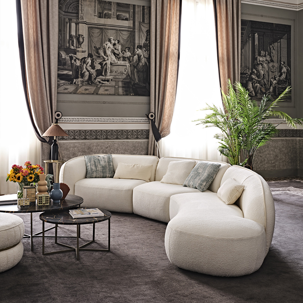 A luxurious living room features a curved cream-colored sofa with multiple cushions, positioned around a black round coffee table adorned with a vase of flowers and decor items. Tall windows with beige and brown curtains, a large potted plant, and classical paintings enhance the elegant ambiance, perfect for easing any blue Monday.