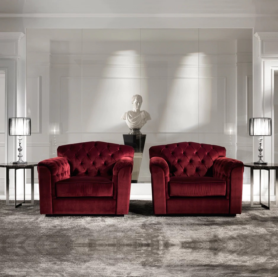 A luxurious living room features two plush, red velvet armchairs with button-tufted backs facing forward, adding pops of colour to the space. They are flanked by two black side tables with elegant table lamps. A white marble bust on a pedestal is placed between the armchairs, against a white paneled wall.