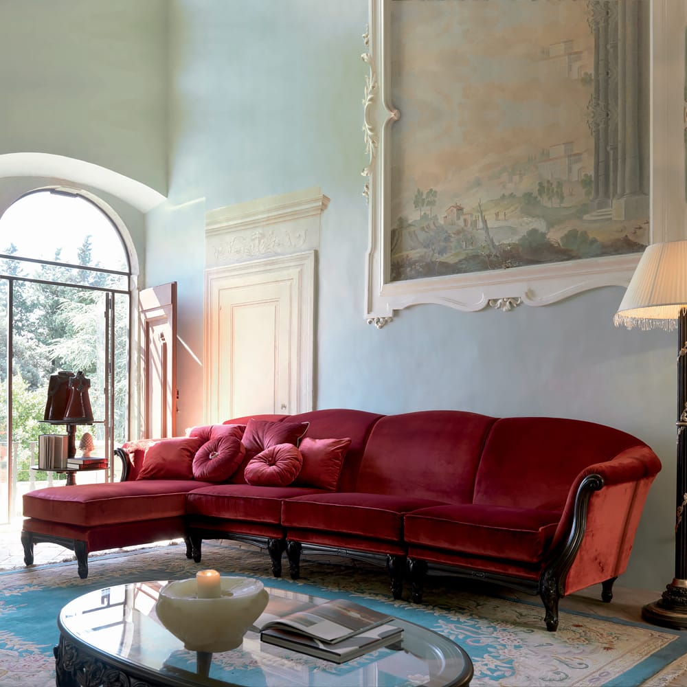 A luxurious living room features a plush red velvet sectional sofa with delicate tufted pillows. Pops of colour enhance the space, complemented by a large arched window with a scenic outdoor view, an elegant wall painting, a stylish floor lamp, and a chic coffee table holding a candle and magazines.