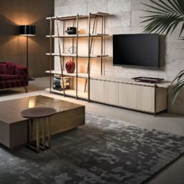 Modern living room featuring a wall-mounted flat-screen TV, bookshelf with decorative items, a wooden storage cabinet, a maroon upholstered couch, a brass and wood coffee table, and a textured rug. A large plant adds greenery, and soft lighting creates a cozy ambiance.