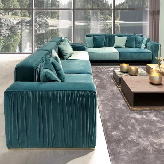 A spacious living room with large floor-to-ceiling windows showcasing a scenic lake view. The room features an L-shaped teal velvet sectional sofa with multiple cushions, a rectangular wooden coffee table with gold decor items, and a gray area rug adding subtle pops of colour.