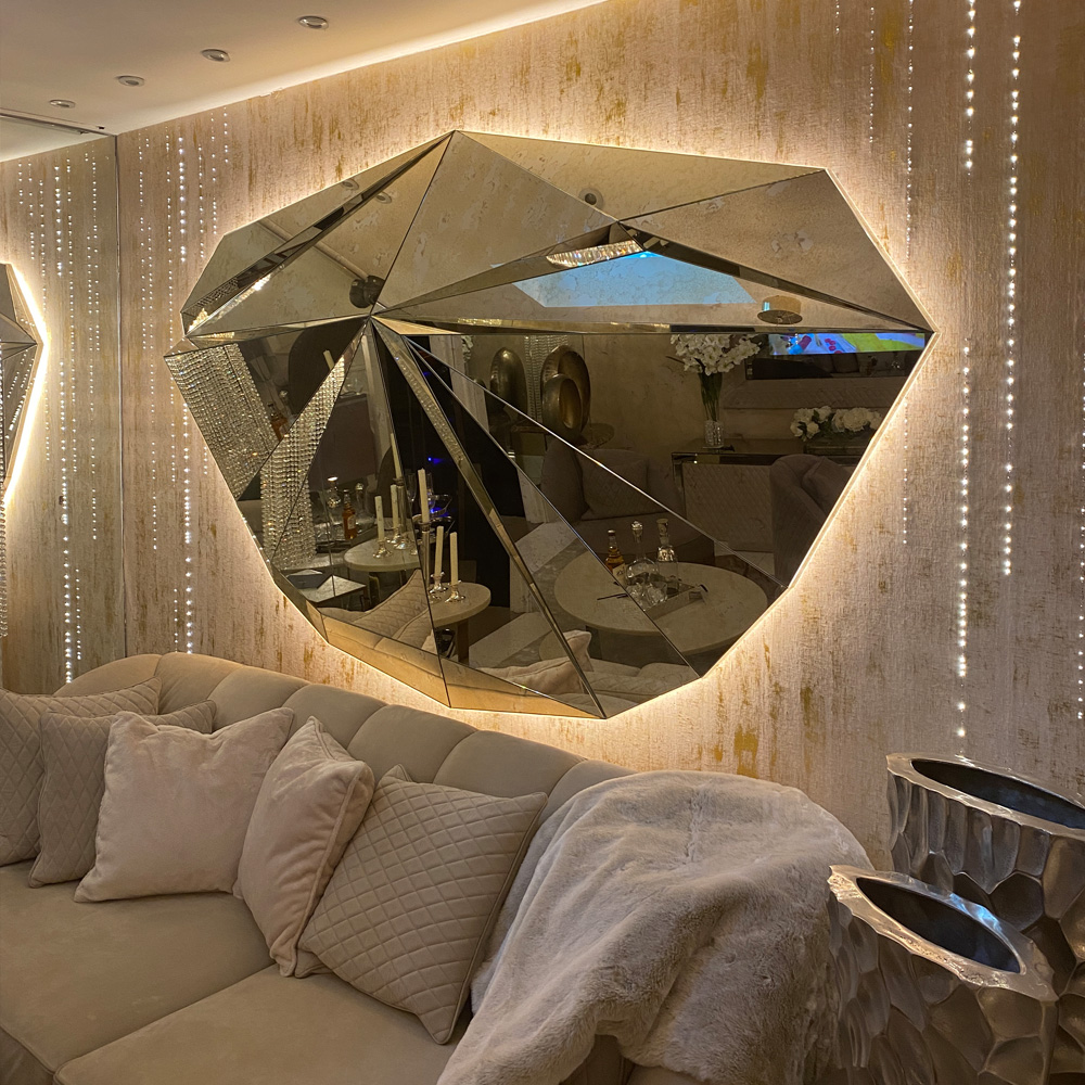 A luxurious living room features a textured wall showcasing the Large Faceted Antique Glass Mirror With Backlight, which beautifully reflects the room's decor. A plush beige sofa is adorned with matching pillows and a throw blanket, while elegant candles and vases are also visible in the reflection.