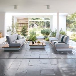 A modern outdoor lounge area with minimalist design, nestled within luxury gardens. It features low, gray cushioned seating with wooden accents arranged around a central low coffee table. The space is open, bright, and surrounded by greenery, with large windows allowing natural light in.