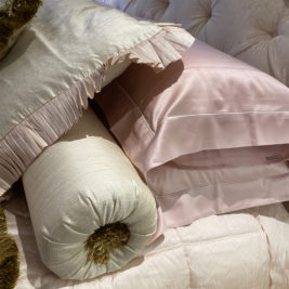 A close-up of a bed featuring luxurious, cream-colored pillows and a cylindrical bolster pillow with fringed ends. The bed is covered with a soft, tufted blanket in a matching shade, accompanied by a light pink, satin pillowcase—perfect for a cozy Valentine's Day retreat.