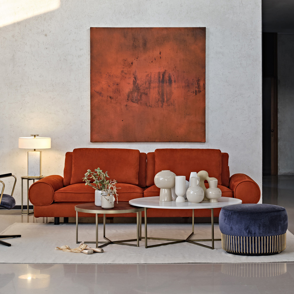 A modern living room with a burnt orange sofa, an abstract painting in matching earthy tones on the wall behind it, two round coffee tables with various vases, a floor lamp, and a blue pouf with golden accents on a light-colored rug.