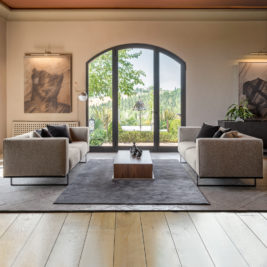 Contemporary living room with two beige sofas facing each other, separated by a wooden coffee table on a gray rug. Large arched window with a view of a lush garden, flanked by modern floor lamps. Abstract art pieces and potted plants enhance the earthy tones of the space.