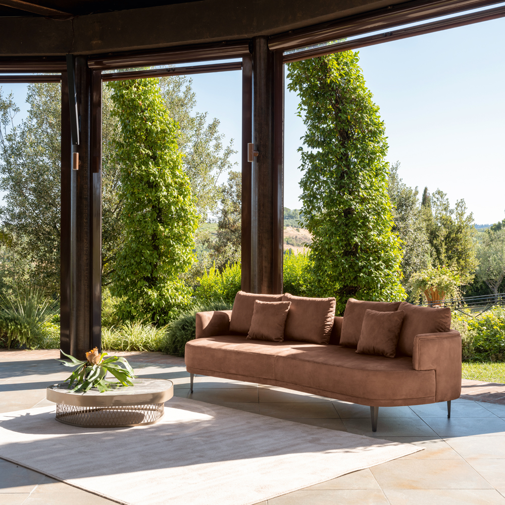 An open-air living space features a brown suede sectional sofa with matching cushions, facing a low round coffee table holding a small plant. The earthy tones of the furniture blend seamlessly with the lush greenery and tall trees surrounding the area, creating a serene and refreshing atmosphere.