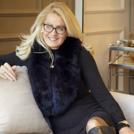 A woman with long blonde hair and glasses sits comfortably on a couch, smiling. She wears a black dress and a dark fur scarf, with one arm resting on the couch and the other on her knee. Behind her, in celebration of International Women's Day, is a bar cart in a warmly lit, elegantly decorated room.