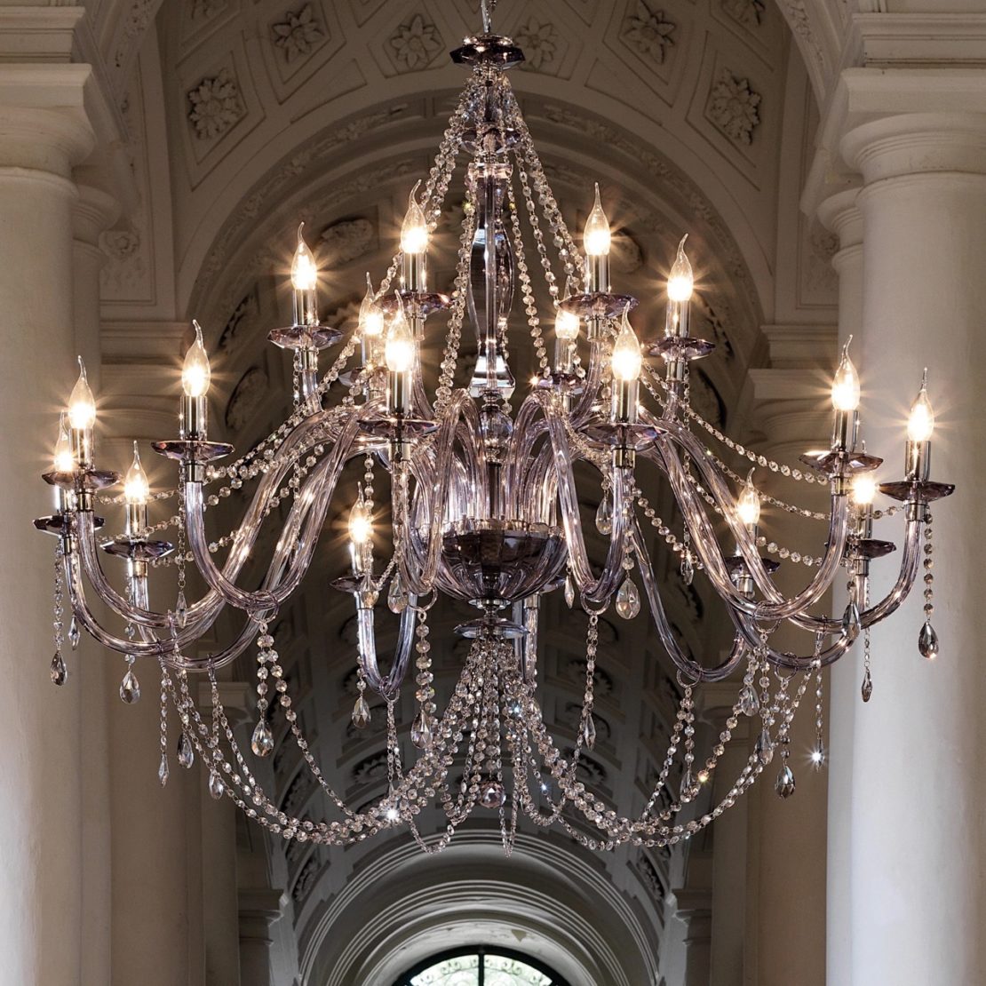 Elegant crystal chandelier with numerous candle-like lights hanging from a vaulted ceiling adorned with intricate moldings. The designer luxury lighting fixture features multiple tiers of crystal strands and pendants, creating a luxurious and grand ambiance.