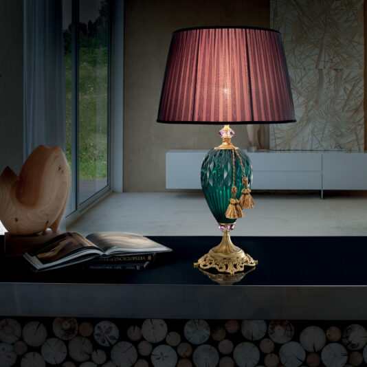 Diamond Faceted Green Glass Table Lamp A stylish room with a Diamond Faceted Green Glass Table Lamp featuring a maroon shade placed on a dark countertop. The lamp sits next to an open book and decorative wooden sculptures, while the background includes a large window with a leafy view and modern decor elements.