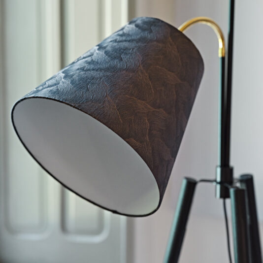 A close-up shot of the Large Modern Studio Style Floor Lamp featuring a textured dark grey lampshade, angled slightly downward. The lamp's gold neck and tripod base are prominently displayed. In the background, a softly lit window is out of focus, adding a touch of natural light to the scene.