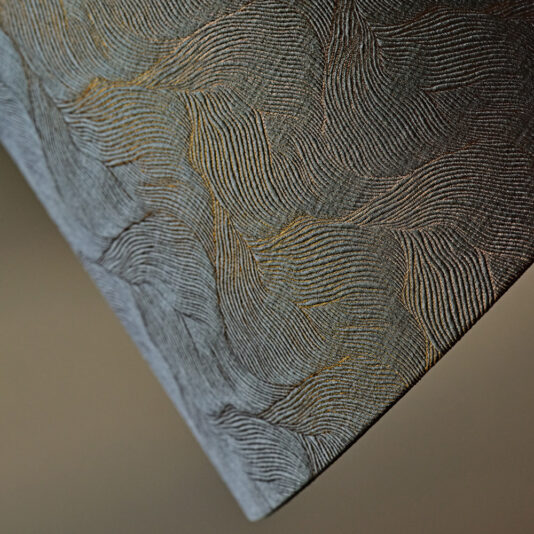 Close-up of a textured surface with swirling, wave-like patterns in shades of grey and black. The design consists of fine, parallel lines creating an abstract, intricate appearance. In the background, the Large Modern Studio Style Floor Lamp casts understated illumination, emphasizing the texture and depth.