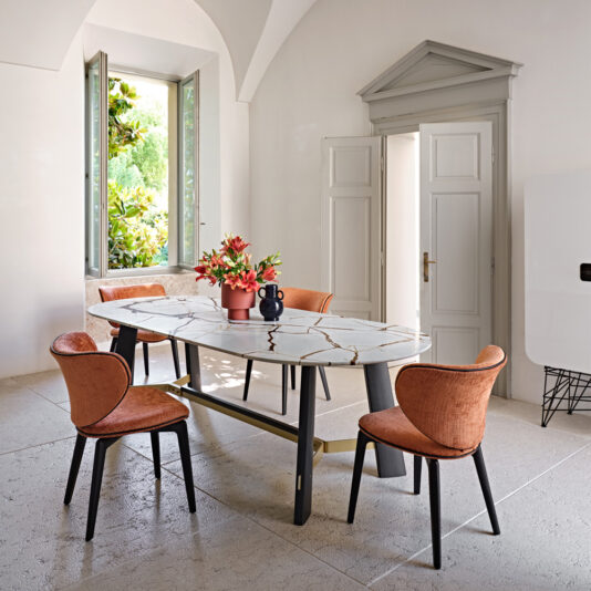 Mid Century Style Dining Chair A bright dining room featuring a white, rectangular marble table adorned with an arrangement of red flowers in vases. Four Mid Century Style Dining Chairs in orange surround the table. The room also has an open window with a view of greenery outside and a partially open arched door in the background.