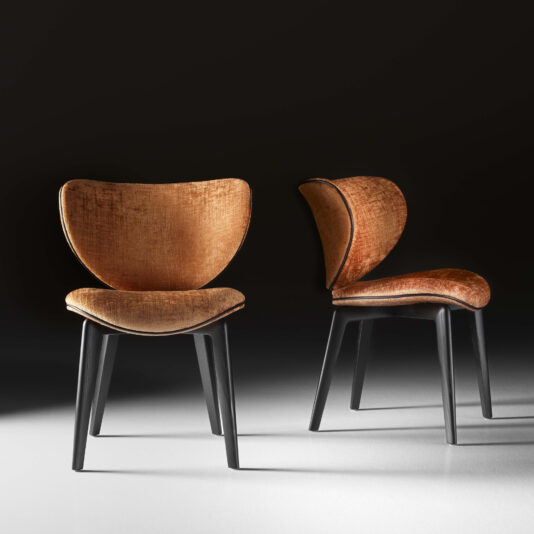 Mid Century Style Dining Chair A pair of Mid Century Style Dining Chairs, featuring modern and stylish designs, with dark wooden legs and plush, golden-brown upholstered seats and backs. One chair is displayed from the front view while the other showcases a side profile, all set against a shadowed, minimalist background.
