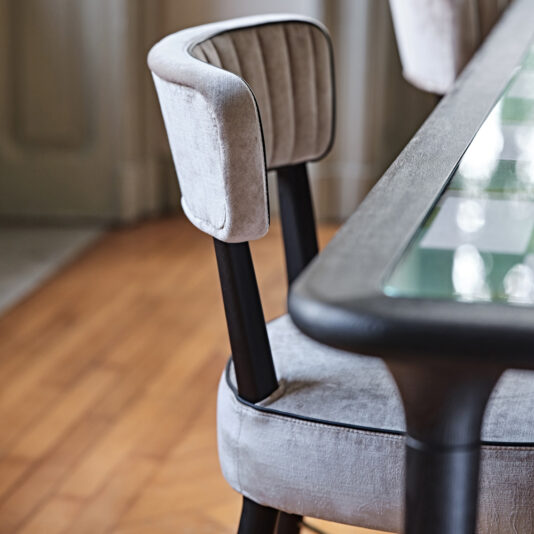 The Modern Designer Velvet Dining Chair, featuring soft gray velvet upholstery and black legs, is artfully positioned beside a glass-top table. The chair boasts a curved backrest with vertical stitching, all set against the backdrop of wooden flooring and a partially visible door.