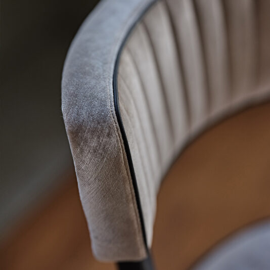 Close-up of a Modern Designer Velvet Dining Chair with a curved backrest. The chair's fabric is a textured, soft grey velvet with vertical stitching details. The edge is trimmed with a thin black line, and the background is softly blurred.