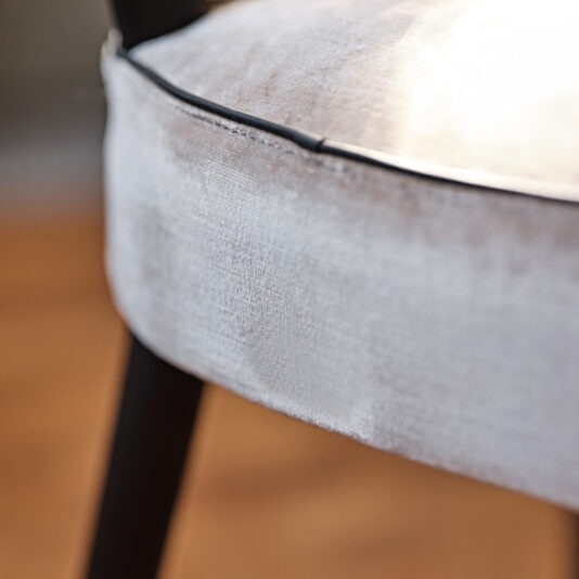Close-up of the Modern Designer Velvet Dining Chair, showcasing its light gray velvet upholstery with black piping detail along the edge. The chair's black legs are partially visible, set against a softly blurred background that accentuates the sleek dining chair's design.