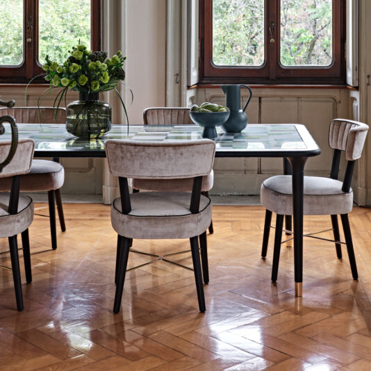 A dining room with a glass-topped table, surrounded by five Modern Designer Velvet Dining Chairs with wooden legs. The table features a dark vase with green flowers, bowls with green apples, and decorative blue vases. Large windows offer a view of lush greenery outside.