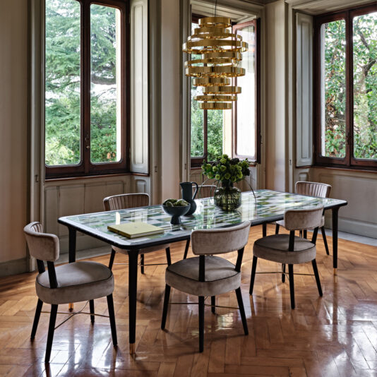 A dining room with a modern rectangular table set for six, featuring a glass top adorned with a green checkered pattern. Surrounding the table are six Modern Designer Velvet Dining Chairs in gray, each cushioned for comfort. Above, a gold-tiered chandelier adds elegance while large windows reveal views of trees outside, allowing natural light to enhance the room's modern designer aesthetic.