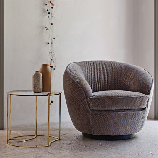 A cozy Modern Luxury Swivel Armchair in gray is positioned next to a round side table with a gold frame and a marble top. The table holds two decorative vases, one brown and one gray. The wall behind features an artistic string and bead decoration, creating a minimalist and modern setting.