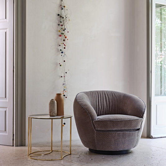 Modern Luxury Swivel Armchair A cozy, rounded Modern Luxury Swivel Armchair in gray is positioned next to a small, elegant side table with a gold frame and glass top. On the table are two decorative vases. The background features a minimalist wall with a string of multicolored ornaments hanging vertically, exuding modern luxury.