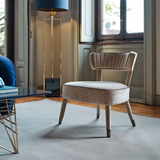 A stylish room featuring the Modern Velvet Occasional Armchair with wooden legs and brass leg caps. The chair is placed on a light gray rug near a modern glass-top coffee table. The room includes a tall floor lamp with a blue shade and large windows with white trim.
