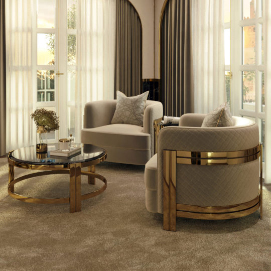 A sophisticated living room features two Modern Curved Armchairs With Brass Frames, a round gold and glass coffee table, a book, a vase with flowers, and two large windows draped with sheer and blackout curtains. The room is bathed in natural light.