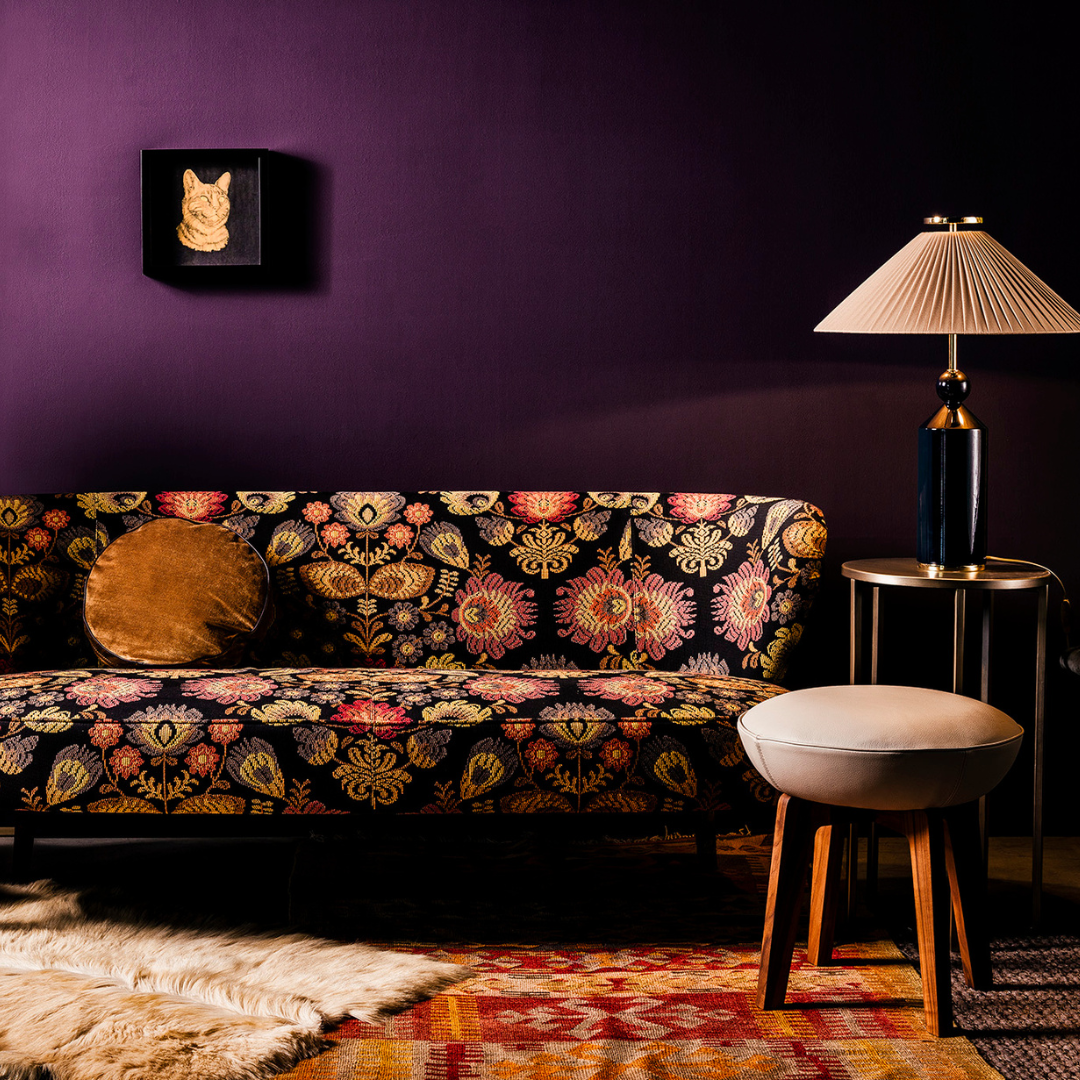 A stylish living room with a dark purple wall, a floral-patterned sofa with a round cushion, and Victorian decor touches such as a small side table with an ornate lamp. A white stool, furry rug, and framed artwork of a cat complete the warmly lit space, creating a cozy ambiance.