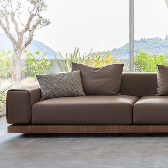 A Contemporary High End Modular Corner Sofa, upholstered in elegant brown leather with a sleek wooden base, graces a bright room filled with natural light from large windows. The chic design showcases two spacious back cushions and a smaller decorative pillow for added style. Beyond the windows, lush green foliage and rolling hills stretch under a brilliant sky.