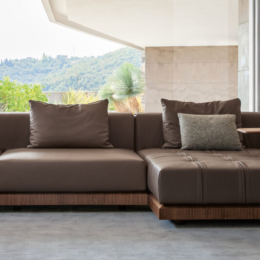 The modern, spacious living room features a Contemporary High End Modular Corner Sofa, adorned with cushions. This L-shaped brown leather sofa enhances the room, which includes an open wall offering a view of green hills and trees, providing a relaxing and airy atmosphere. The light gray flooring complements the elegant decor.