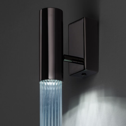 The Contemporary Vertical Glass Tube Wall Light boasts a sleek, modern design with a cylindrical black metal casing and a clear ribbed glass tube beneath it, beautifully casting soft white light onto the gray wall behind it.