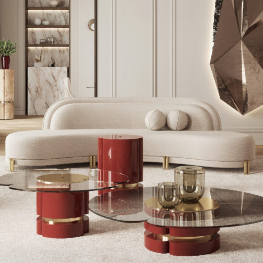 A modern living room features a curved beige sofa with two round cushions, reminiscent of jellyfish. In front of it, two glass tables with red and gold cylindrical bases add a splash of color. A large geometric wall art piece is displayed on the right, while sleek shelves and cabinets enhance the interior design.