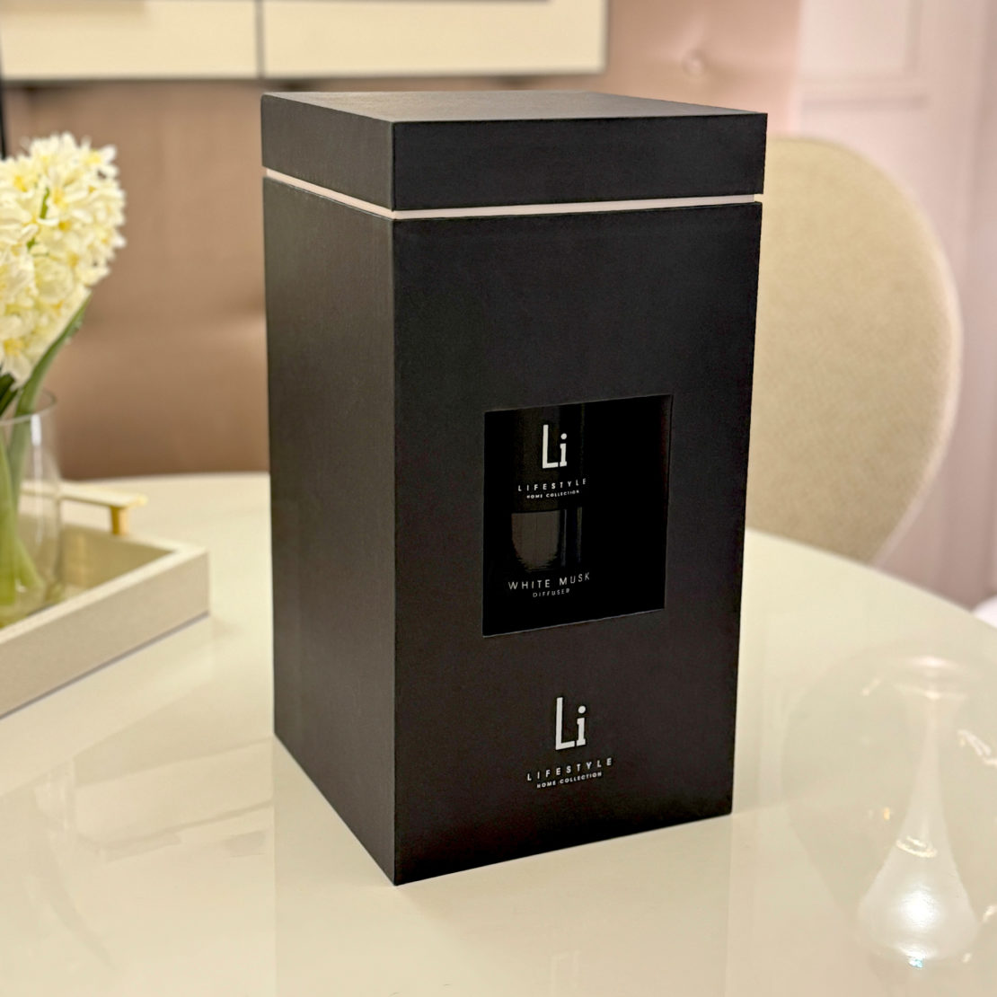 A black rectangular box labeled "Li" and "White Musk" with a small window displaying the luxury home fragrance inside is placed on a white table. In the background, there is a white cushioned chair, some flowers in a vase, and a golden tray with items on it.