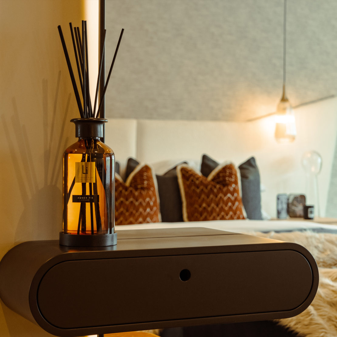 A close-up of a modern bedside shelf with an Extra Large Luxury Amber Fie Room Diffuser gracing it. In the background, a cozy bed adorned with patterned throw pillows and illuminated by a hanging pendant light creates a warm and inviting bedroom atmosphere.