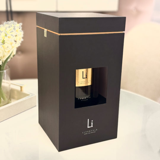 A sleek black box with gold accents, showcasing a transparent window that reveals the Extra Large Luxury Amber Fie Room Diffuser inside. The box is branded "Lifestyle" with a minimalist "Li" design. It sits on a white table beside a tray holding a vase of white flowers and a book.