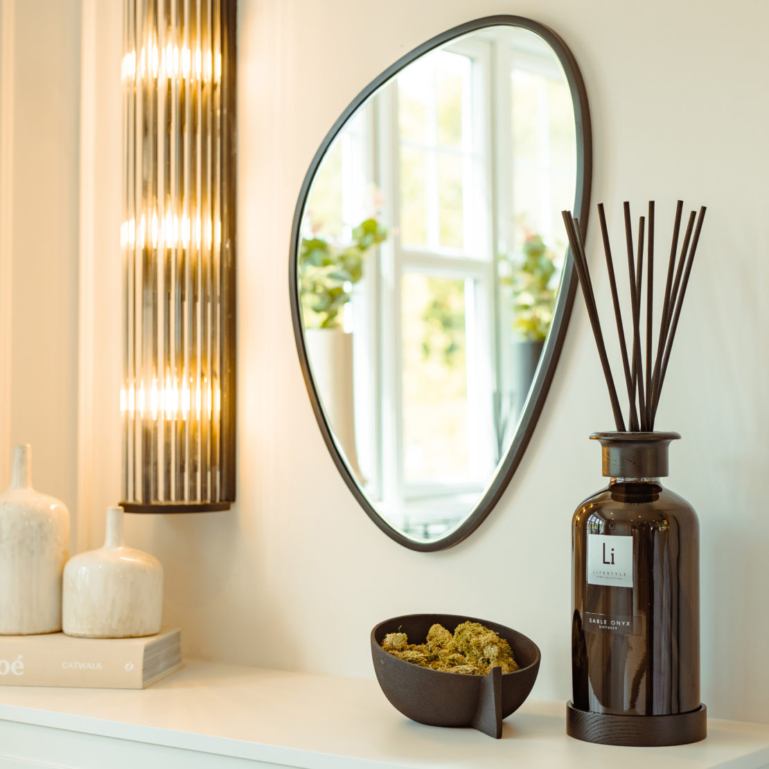 A modern living space showcases a teardrop-shaped mirror on the wall, a tall, sleek lamp emitting warm light, ceramic vases, and a decorative bowl filled with green moss. Featured prominently on a white shelf is the Extra Large Luxury Sable Onyx Room Diffuser. In the background, you can see plants by a bright window.