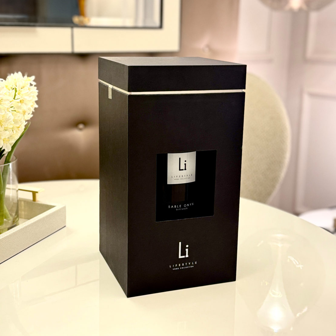 A sleek black box adorned with the logo and name "Li" on the front suggests an Extra Large Luxury Sable Onyx Room Diffuser. The box is elegantly positioned on a round table beside a vase of white flowers. In the background, a cushioned chair, mirror, and light-colored wall enhance the setting's modern sophistication.