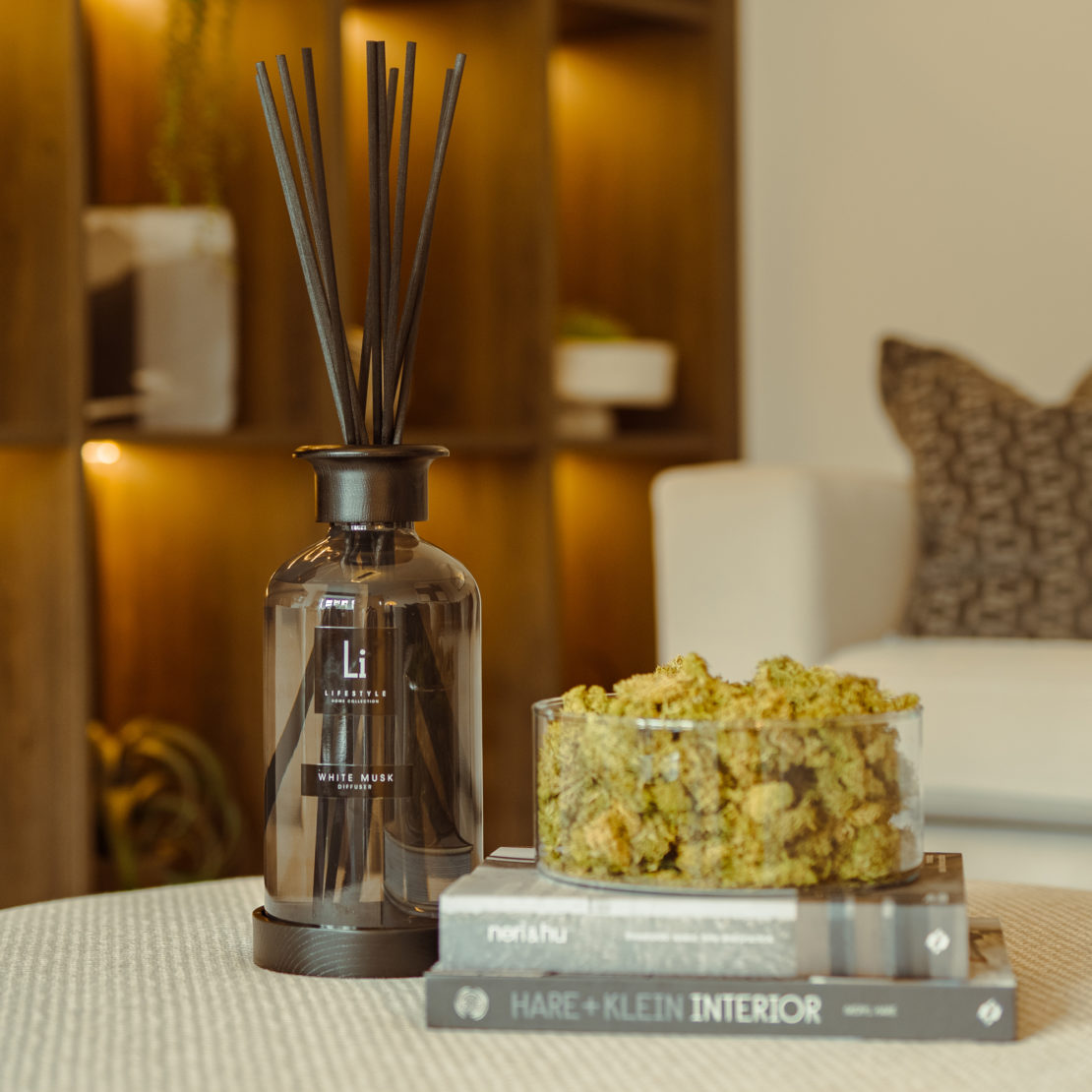 A cozy living room scene features the Extra Large Luxury White Musk Room Diffuser with dark sticks on a circular wooden tray atop a stack of three interior design books. Beside the diffuser, there is a transparent bowl filled with green dried moss. In the background is a comfortable white sofa.