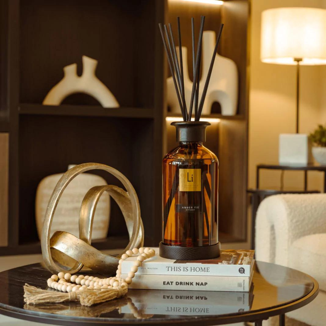 An amber diffuser bottle with reed sticks, offering a touch of luxury home fragrance, is placed on a glass coffee table alongside books titled "EAT DRINK NAP" and "THIS IS HOME." Beside the books is a decorative golden object and a beaded garland. The background features a warm, cozy living room setup.