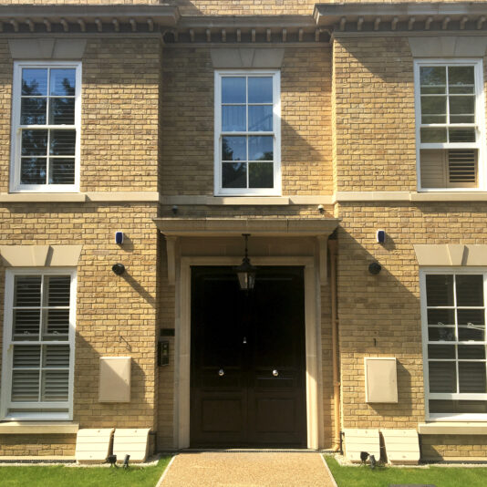 A symmetrical brick building facade featuring a central double door with a lantern light above it. The door is flanked by two sets of windows on each side, and there are two additional windows directly above. The well-kept lawn adds to its charm, highlighting the meticulous design stage.