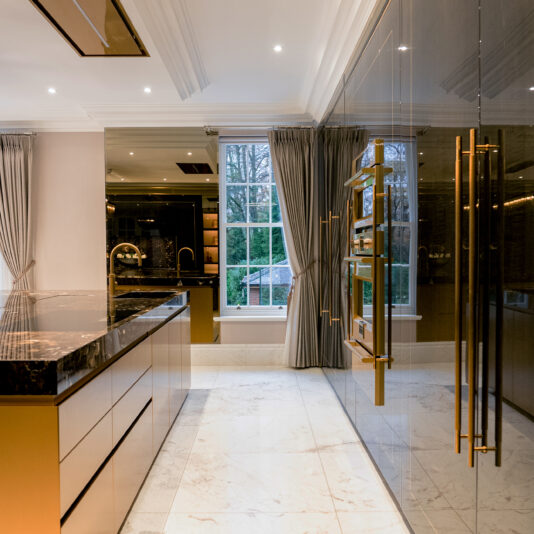 A luxury family kitchen with a marble island countertop, beige cabinets, and large windows. The room features glossy reflective surface cabinets with gold handles and built-in appliances. Grey curtains frame the windows, while recessed ceiling lights provide illumination, creating a warm and inviting space.