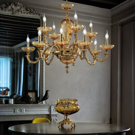 A luxurious dining room featuring a Large Hand Blown Amber Glass Chandelier suspended above a round black table, which holds a decorative gold vase. In the background, a large framed mirror reflects part of the room, and a mantel adorned with decorative items is visible.