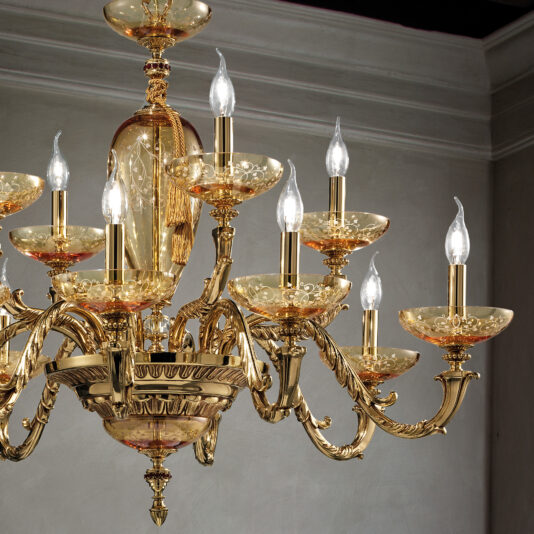The Large Hand Blown Amber Glass Chandelier features numerous ornate, golden arms, each adorned with glowing candle-like light bulbs. The large hand blown amber glass elements add a luxurious and elegant touch to the intricate designs. When set against a plain, light-colored wall, it becomes a stunning focal point in the room.