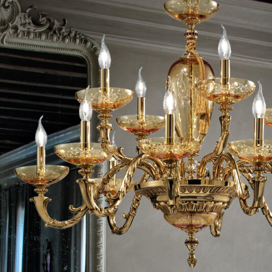 A luxurious gold chandelier with multiple ornate arms and candle-like light bulbs is seen hanging prominently. The background showcases an elegant mirror with an intricate frame, reflecting the Large Hand Blown Amber Glass Chandelier and adding to the opulent ambiance of the room.
