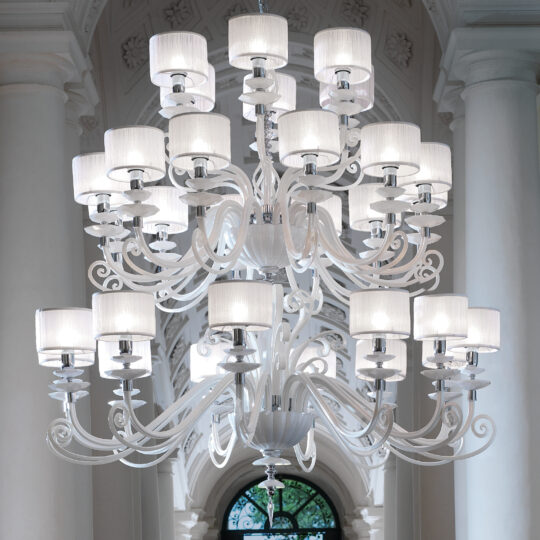 A grand, multi-tiered chandelier with numerous white lampshades and ornate white metalwork hangs from the ceiling of a room adorned with tall, white columns and arched windows. The Large White Glass Modern Chandelier exudes elegance and sophistication.