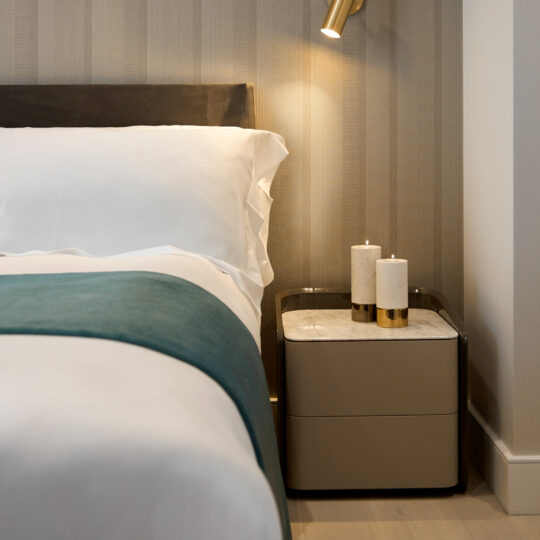 A modern bedroom features a bed adorned with white linens and a teal blanket elegantly draped over the edge. Beside it stands the Contemporary Bedside Cabinet with Drawers, supporting two marble and gold cylindrical candles. A wall-mounted lamp casts a warm glow above, illuminating the neutral, striped wallpaper in the background.