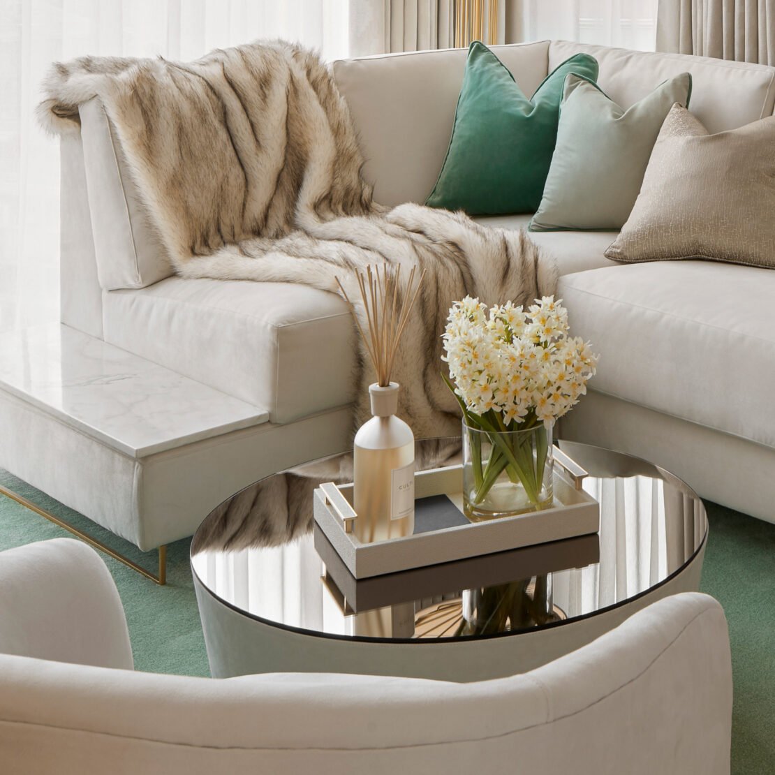 A modern living room showcases a high-end corner sofa bed with an integrated side table, adorned with a faux fur throw and green and beige pillows. A circular coffee table with a mirrored surface holds a tray of decorative items and a vase of white flowers. Beige curtains, along with an elegant additional side table and green rug, complete the space. The finishing touches make the room decor