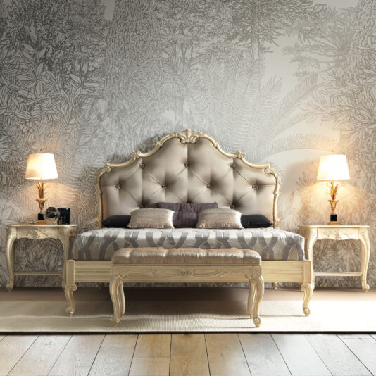 A luxurious bedroom showcases the Romantic Button Upholstered Bed, complete with an ornate tufted headboard and patterned bedding. Two side tables, each holding a lamp that casts a warm glow, flank the bed. The wallpaper behind features a detailed, nature-inspired design.
