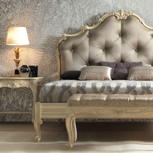 A luxurious bedroom features the Romantic Button Upholstered Bed, adorned with an ornate beige headboard. The romantic bed is dressed with patterned pillows and a matching bedspread. Beside the bed sits a nightstand complete with a decorative table lamp and various accessories. The wallpaper showcases a subtle leaf-patterned design.